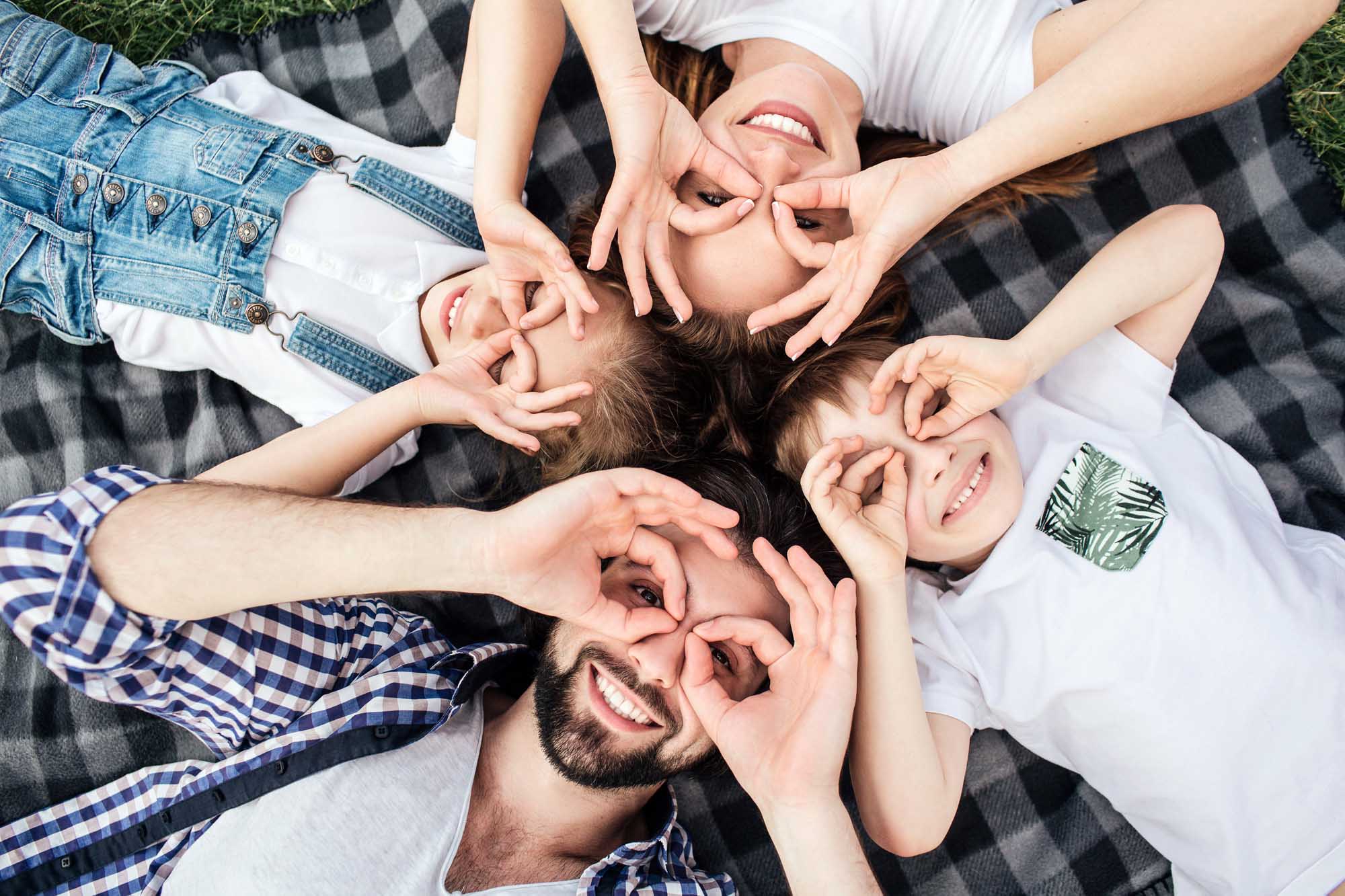 Funny picture of family doing funny rounds with their fingers on eyes. They are playing. All of them are lying on blanket and smiling. They look happy.